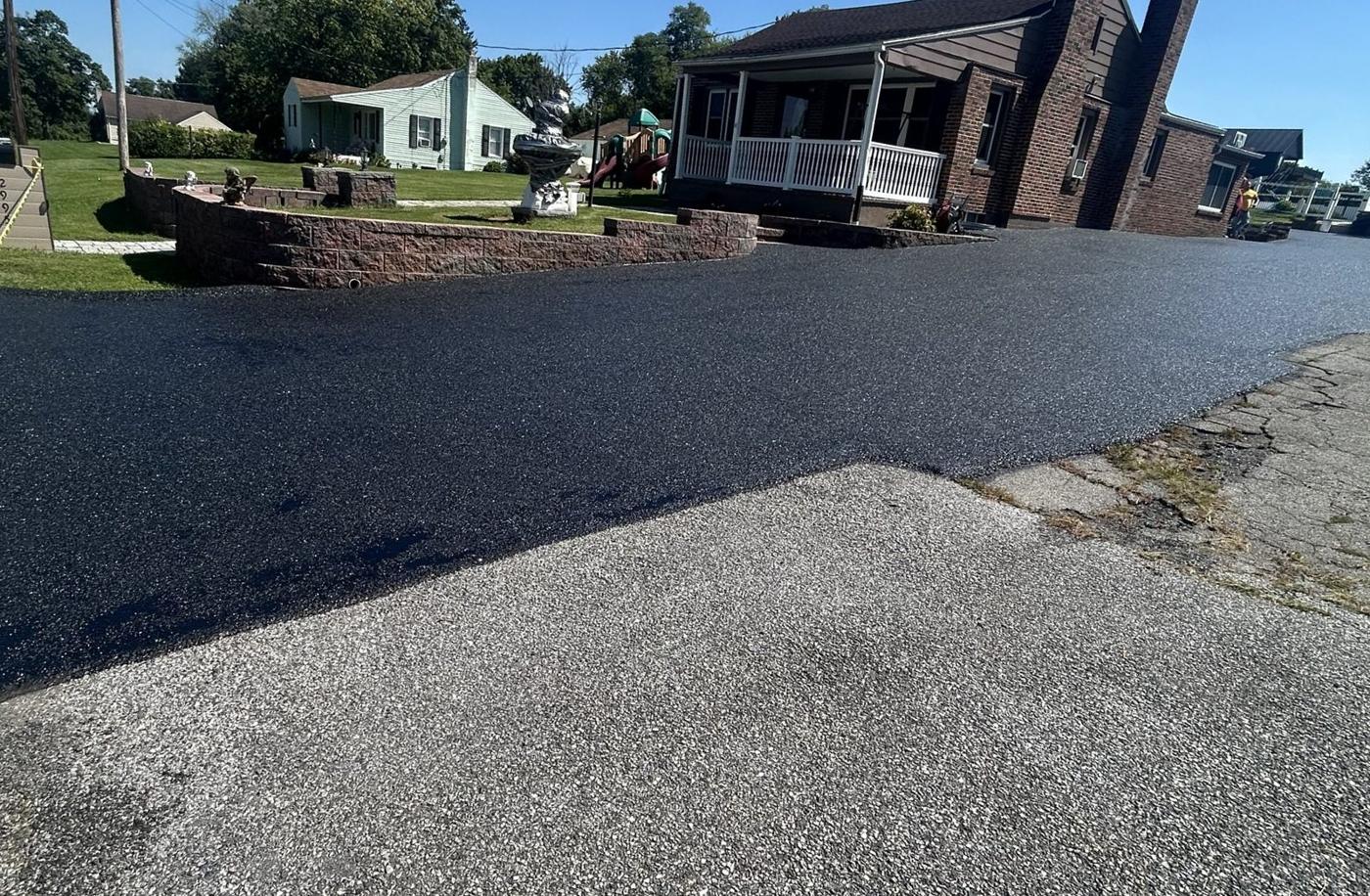 A freshly paved black asphalt driveway beside a brick house, finished by All About Asphalt